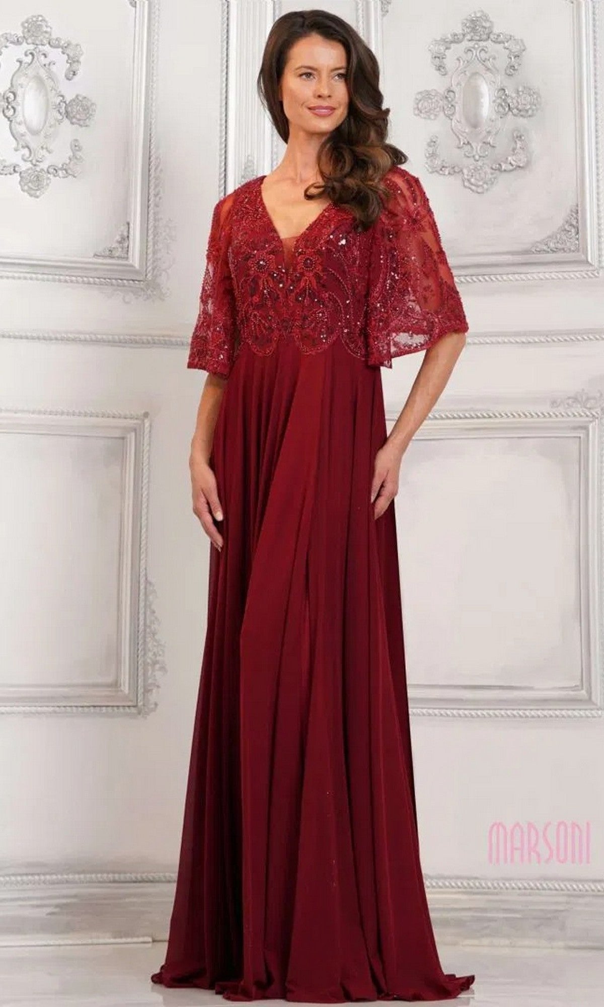 A woman in the Marsoni MV1302 Long Formal Dress, a floor-length deep red beaded gown with lace overlay and short sleeves, stands against an ornate white wall.
